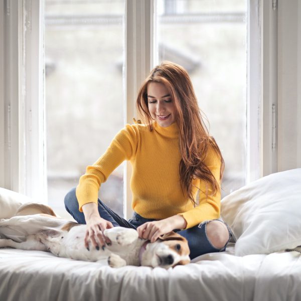 Photo of Smiling Woman in a Yellow Turtleneck Sweater Sitting on Bed Petting Her Dog